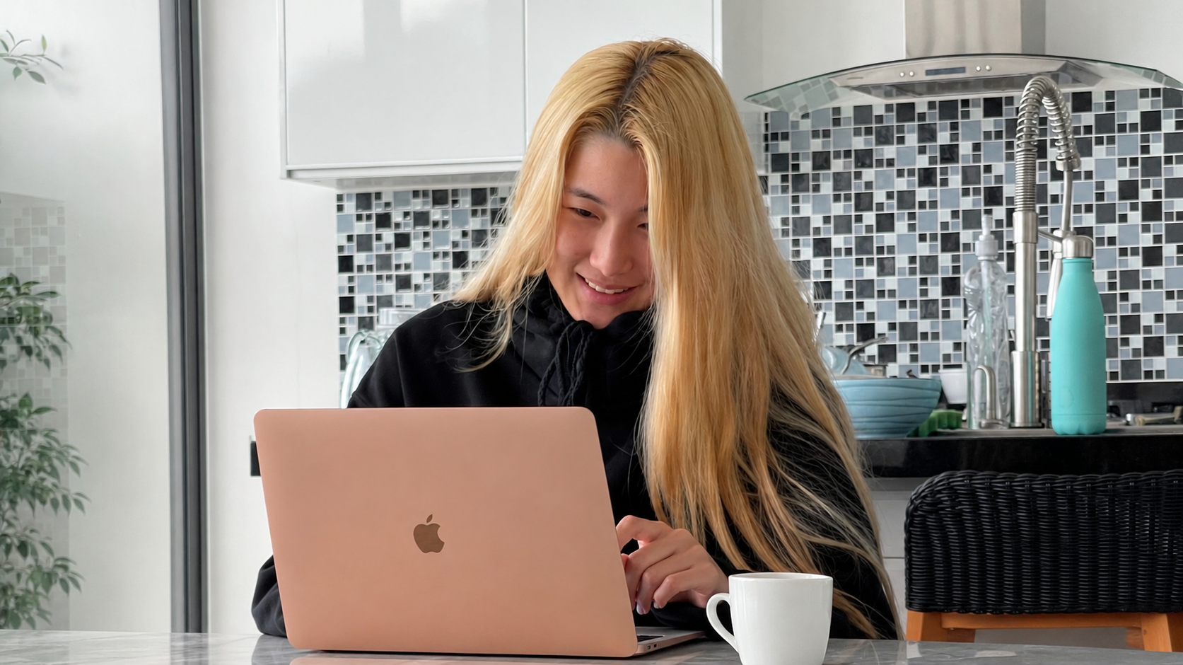 Cathy at a kitchen counter, long blonde hair, rose-gold MacBook open, white mug beside her